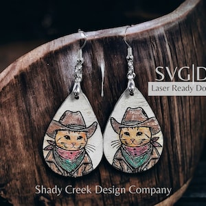 May include: A pair of laser-cut wooden earrings featuring a cartoon cat wearing a cowboy hat and bandana. The earrings are shaped like teardrops and have silver hooks. The text "SVG|DXF Laser Ready Download" is visible in the image.