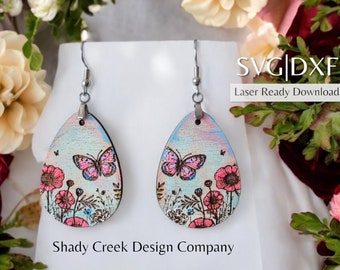May include: Teardrop earrings with a butterfly and floral design. The earrings have a light blue and pink watercolour background, a pink butterfly, and pink flowers. The text "SVG|DXF Laser Ready Download" and "Shady Creek Design Company" are visible.