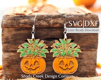 Puede incluir: Un par de pendientes con temática de Halloween. Cada pendiente presenta una calabaza naranja tallada con una cara sonriente, coronada con hojas verdes. Los pendientes cuelgan de ganchos plateados. El texto "SVG|DXF Laser Ready Download" es visible.