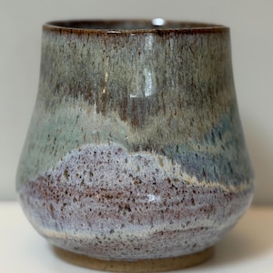 Op de afbeelding: Een handgemaakte keramische beker met een uniek, gelaagd glazuur. De kleuren gaan over van een diepbruine rand naar tinten groen, blauw en paars, met een textuur, gespikkelde uitstraling. De basis is een natuurlijke, ongeglazuurde kleikleur.