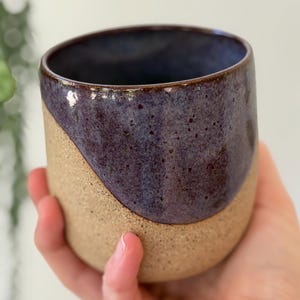 Puede incluir: Un vaso de cerámica hecho a mano con un diseño de dos tonos. La mitad superior es de un morado profundo y moteado, mientras que la mitad inferior es de un beige natural y moteado. La taza tiene un borde liso y curvado.