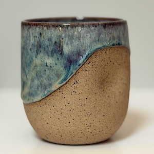 Könnte beinhalten: Eine handgefertigte Keramiktasse mit einem einzigartigen Design. Die obere Hälfte ist in Blautönen glasiert, die in eine gesprenkelte, unglasierte, beige Basis übergehen. Der Rand ist dunkelbraun. Diese Steinzeugtasse ist perfekt für Kaffee oder Tee.