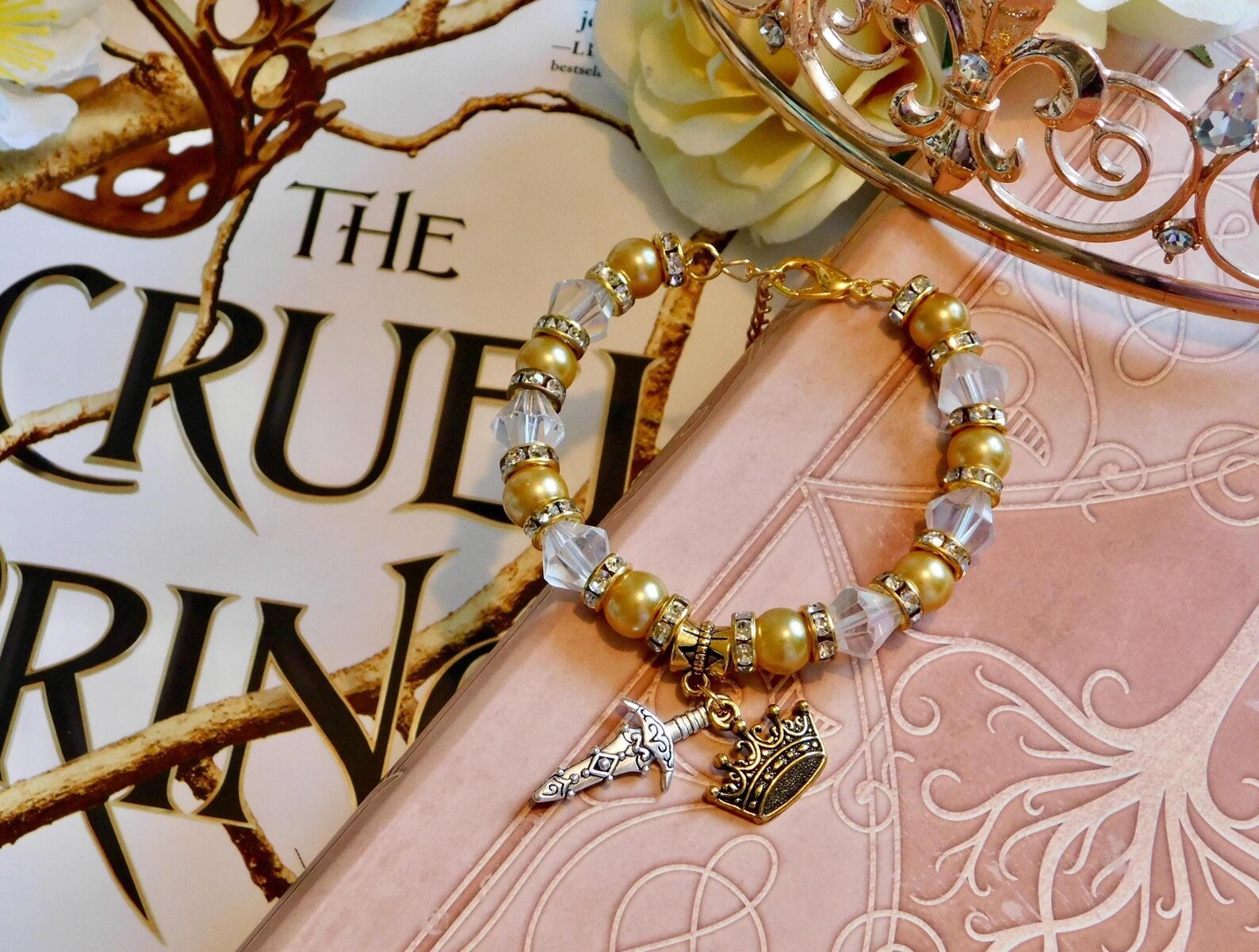 Focused in the center is a gold and clear beaded bracelet with a sword charm and crown charm. Behinf the charm is 'The Cruel Prince' book title.