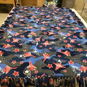May include: A blue and gray fleece tie blanket with a camouflage pattern featuring red airplanes and stars. The blanket has the text "ANGELS 15" and "ST" repeated throughout the pattern.