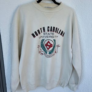 K&ouml;nnte beinhalten: Cremefarbenes Sweatshirt mit den Worten "North Carolina State University" in weinroter und schwarzer Schrift. Das Sweatshirt hat ein Wolfpack-Logo in der Mitte mit einem rot-gr&uuml;nen Emblem. Das Sweatshirt mit Rundhalsausschnitt h&auml;ngt an einem schwarzen Kleiderb&uuml;gel.
