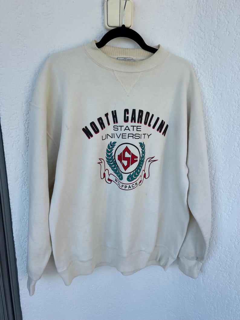 K&ouml;nnte beinhalten: Cremefarbenes Sweatshirt mit den Worten "North Carolina State University" in weinroter und schwarzer Schrift. Das Sweatshirt hat ein Wolfpack-Logo in der Mitte mit einem rot-gr&uuml;nen Emblem. Das Sweatshirt mit Rundhalsausschnitt h&auml;ngt an einem schwarzen Kleiderb&uuml;gel.