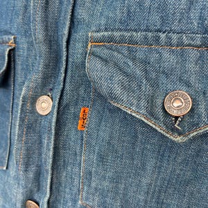 May include: A close-up of a blue denim shirt with a patch that says "Levi's" on the chest. The shirt has a pocket on the left side and several metal buttons.