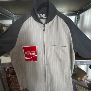 Op de afbeelding: Grijze en zwarte jas met ritssluiting, witte krijtstrepen en een Coca-Cola logo patch. De jas heeft een zwarte kraag en mouwen, en een voorzak. Het Coca-Cola logo is rood en wit.