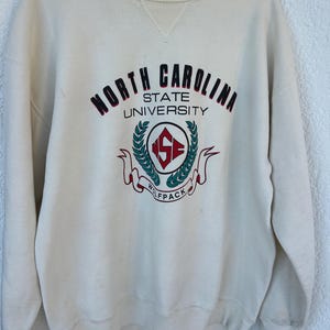 K&ouml;nnte beinhalten: Cremefarbenes Sweatshirt mit den Worten "NORTH CAROLINA STATE UNIVERSITY" in kastanienbraunen und schwarzen Buchstaben. Das Sweatshirt hat ein rundes Emblem mit "NC STATE" und "WOLFPACK" in Rot und Gr&uuml;n.