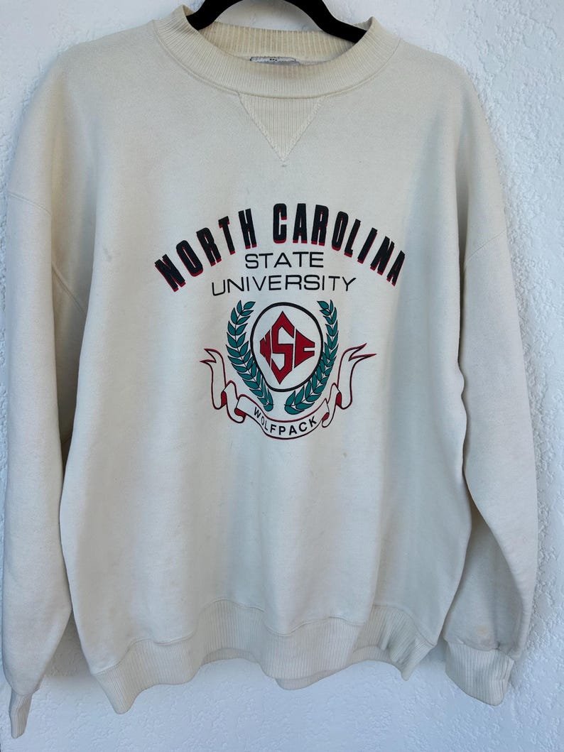 K&ouml;nnte beinhalten: Cremefarbenes Sweatshirt mit den Worten "NORTH CAROLINA STATE UNIVERSITY" in kastanienbraunen und schwarzen Buchstaben. Das Sweatshirt hat ein rundes Emblem mit "NC STATE" und "WOLFPACK" in Rot und Gr&uuml;n.