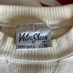 K&ouml;nnte beinhalten: Cremefarbenes Sweatshirt mit geripptem Rundhalsausschnitt und einem aufgen&auml;hten V-f&ouml;rmigen Detail. Das Etikett lautet "Velva Sheen" in marineblauer Schrift, mit "MADE IN U.S.A." und "ADULT XL" darunter.