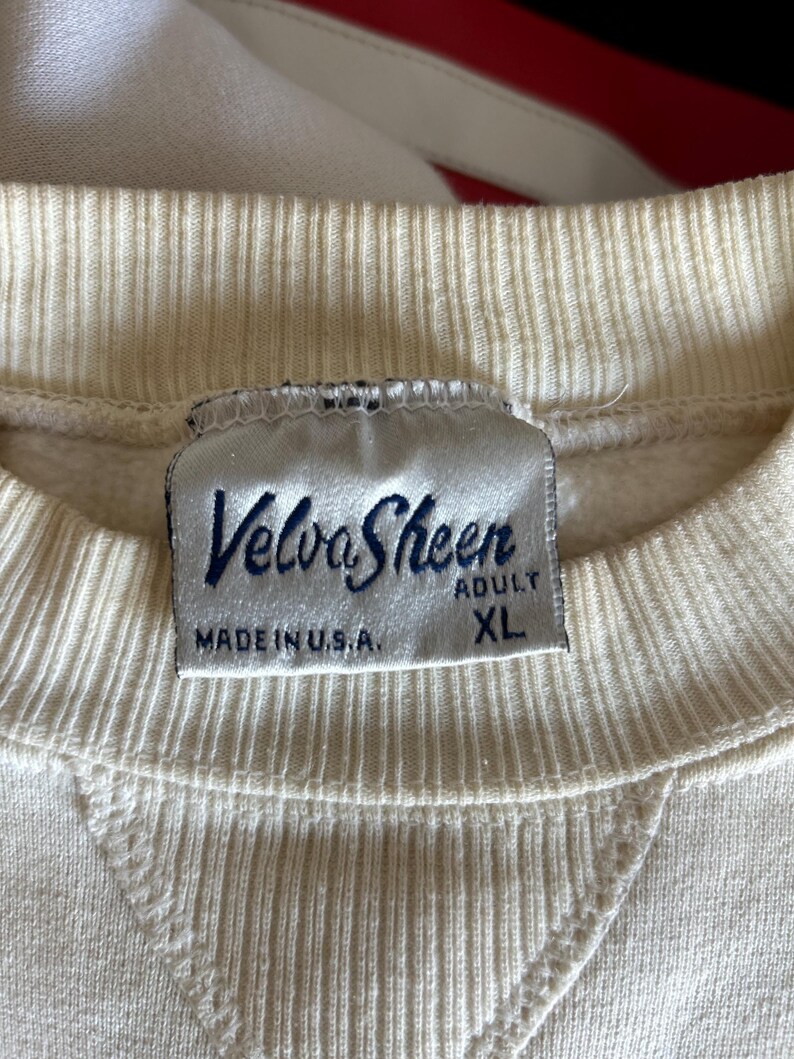 K&ouml;nnte beinhalten: Cremefarbenes Sweatshirt mit geripptem Rundhalsausschnitt und einem aufgen&auml;hten V-f&ouml;rmigen Detail. Das Etikett lautet "Velva Sheen" in marineblauer Schrift, mit "MADE IN U.S.A." und "ADULT XL" darunter.