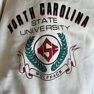 K&ouml;nnte beinhalten: Cremefarbenes Sweatshirt mit dem Text "North Carolina State University" in schwarzer und roter Schrift. Das Design umfasst ein rundes Emblem mit "NSF" und ein "Wolfpack" Banner.