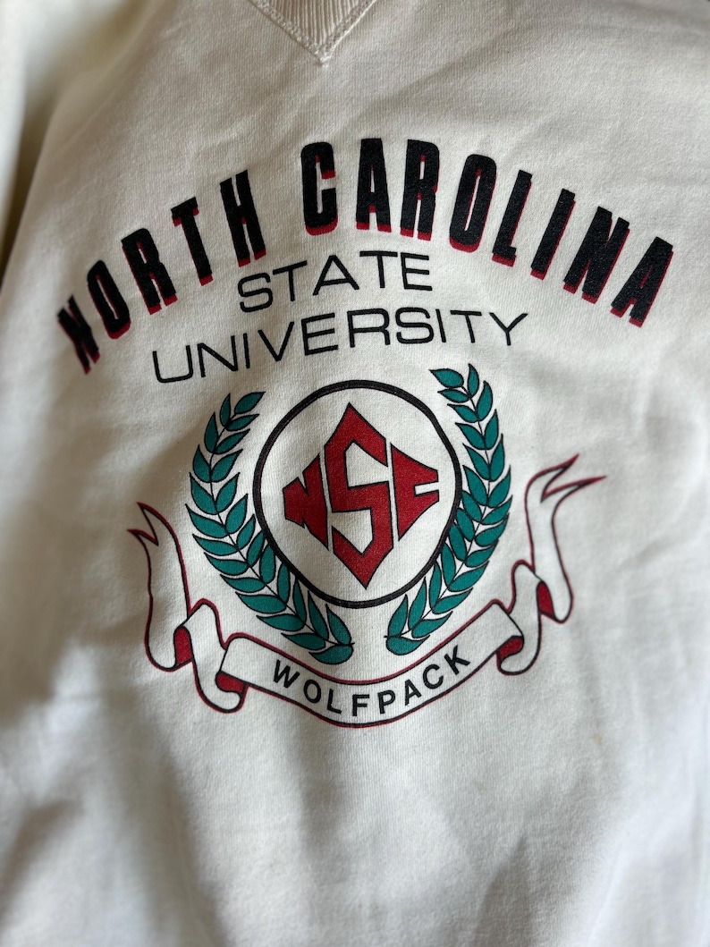K&ouml;nnte beinhalten: Cremefarbenes Sweatshirt mit dem Text "North Carolina State University" in schwarzer und roter Schrift. Das Design umfasst ein rundes Emblem mit "NSF" und ein "Wolfpack" Banner.