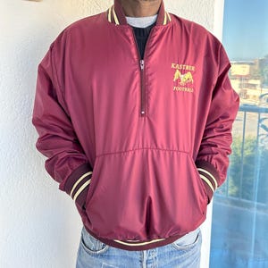 May include: A maroon windbreaker jacket with a zipper closure and a white and gold embroidered logo that says "Kastner Football".