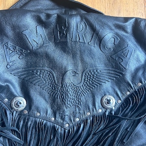 May include: A black leather jacket featuring an embossed eagle design and the word AMERICA across the top. The garment includes silver-toned metal studs, two decorative circular metal buttons, and long black leather fringe along the bottom edge.