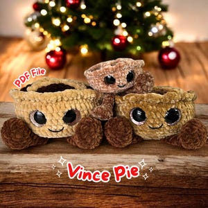 Puede incluir: Tres juguetes de peluche con forma de tarta con caras sonrientes y ojos grandes y brillantes. Las tartas son en tonos marrones y beige, con un relleno más oscuro. El texto "Vince Pie" se muestra en rojo. El fondo incluye un árbol de Navidad.