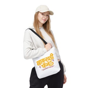 Könnte beinhalten: Weiße Tragetasche mit schwarzem Schultergurt. Die Tasche hat orangefarbenen Text in einer stilisierten Schrift, der "BAKARWADI FAN" und "BAKARWADI FOREVER" in kleinerer Schrift liest. Die Tasche wird von einer Person getragen, die einen cremefarbenen Kapuzenpullover und eine Kappe trägt.