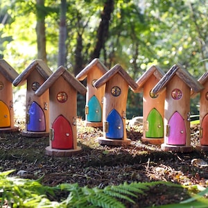 May include: A selection of handcrafted wooden fairy houses with doors in various colours: yellow, purple, red, teal, blue, green, pink, and orange. Each house has a small window and a wooden shingle roof. The houses are set in a natural outdoor environment.