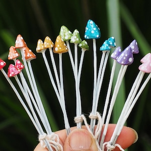 May include: A selection of miniature mushroom decorations in various colours. Each mushroom features a white stem and a cap painted in red, orange, yellow, green, blue, purple, and pink, with white polka dots. The mushrooms are tied together with twine.
