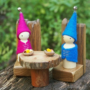 May include: Two wooden gnome figures with felt hats and jackets sit at a small table. The gnome on the left wears a pink hat and jacket, while the one on the right wears blue. A small silver bell tops each hat.