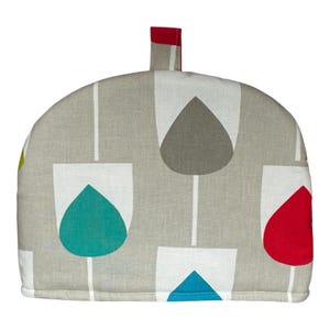 May include: A dome-shaped tea cosy featuring a light beige fabric base with a repeating pattern of white tulip-like shapes containing solid teardrop motifs in teal, red, grey, and blue. A small red fabric loop is attached to the top centre.
