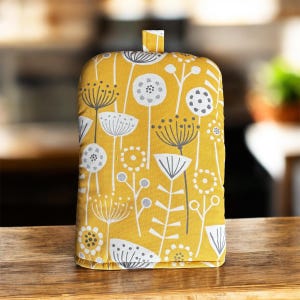 May include: A yellow tea cosy featuring a floral design in white and grey. The cosy has a loop at the top and is designed to fit over a teapot. The fabric has a repeating pattern of stylized flowers and leaves.