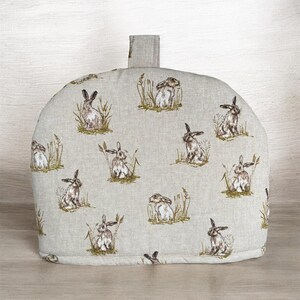 May include: A beige tea cosy featuring a repeating pattern of rabbits amongst grass. The cosy has a rectangular handle and is made from a textured fabric. The design is reminiscent of a rural scene.