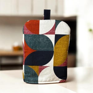 May include: A colourful oven glove with a geometric pattern in shades of red, navy, teal, mustard yellow, and white. The design features semi-circles and lines. A small, dark blue loop is attached at the top for hanging.