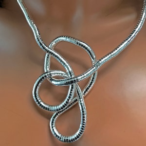 May include: A silver-colored necklace with a unique, knotted design. The necklace is made of a flexible, ribbed material that reflects light, creating a shimmering effect. The design features a series of loops and curves, adding visual interest.