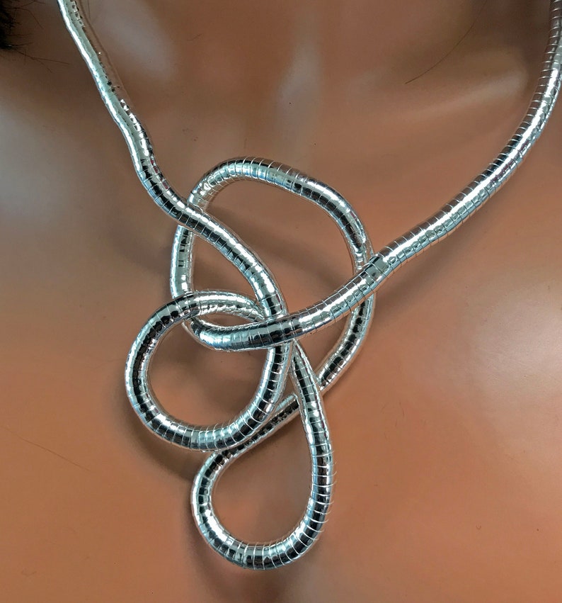 May include: A silver-colored necklace with a unique, knotted design. The necklace is made of a flexible, ribbed material that reflects light, creating a shimmering effect. The design features a series of loops and curves, adding visual interest.