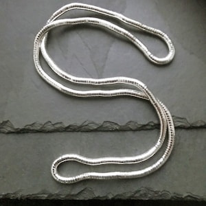 May include: A silver-colored necklace with a snake chain design, lying on a dark gray surface. The necklace is made of multiple small, interconnected links, creating a smooth, flexible appearance. The chain is curved and looped, showcasing its design.