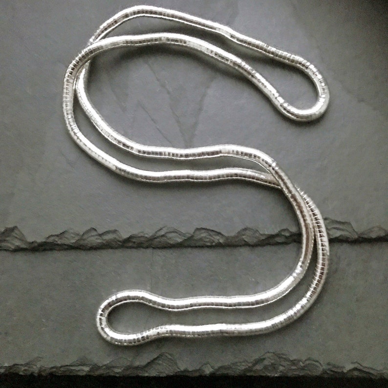 May include: A silver-colored necklace with a snake chain design, lying on a dark gray surface. The necklace is made of multiple small, interconnected links, creating a smooth, flexible appearance. The chain is curved and looped, showcasing its design.