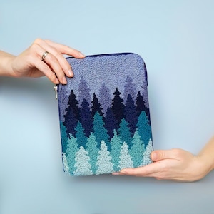 Könnte beinhalten: Eine rechteckige, mit Reißverschluss versehene Tasche mit einem Waldmotiv. Die Tasche hat einen strukturierten, blauen Hintergrund mit einem Muster aus immergrünen Bäumen in Türkis-, Marine- und Indigotönen. Der Reißverschluss ist silberfarben.