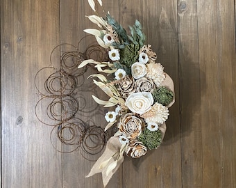 May include: A rustic wreath made from a metal spring with a variety of dried flowers and greenery. The flowers are white and cream colored, and the greenery is a light green. The wreath is tied with a burlap ribbon.