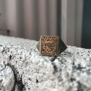 May include: A square-shaped signet ring with a gold-coloured band and a black inscription in Arabic script.