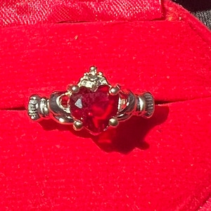 May include: A gold ring with a red heart-shaped gemstone and a crown design. The ring is set in a red velvet box.