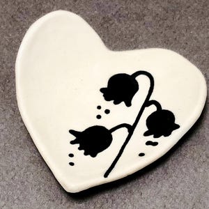 May include: A heart-shaped, off-white ceramic dish. The dish features a black floral design with three bell-shaped flowers and small dots. The design is centered on the dish, which has a slightly raised edge.