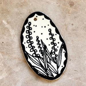 May include: Oval-shaped, white pendant with a black floral design. The pendant features a black border with a scalloped edge and a small hole at the top. The design includes stylized leaves and dotted stems.