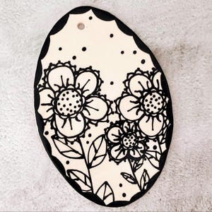 May include: A cream-colored, oval-shaped ceramic piece with a black scalloped border. The design features black floral illustrations with dotted centers and leaves. Small black dots are scattered across the surface. A small hole is at the top.