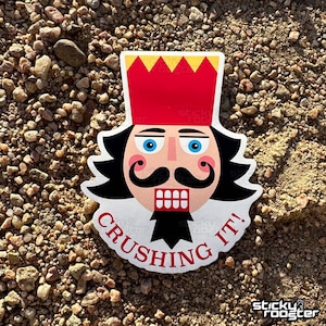 May include: A white sticker with a cartoon illustration of a nutcracker with a red hat and a black mustache. The text "CRUSHING IT!" is written in red around the nutcracker.