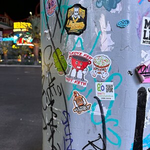 Smoke Meat Not Meth sticker with a little grill giving a thumbs up. Sticker is on a pole with other stickers.