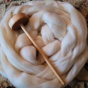 May include: A wooden drop spindle with a light-colored shaft and a round, flat whorl rests on a large pile of white wool roving. The wool is arranged in a circular pattern, creating a soft, textured appearance. The spindle is ready for spinning.