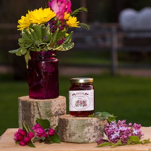 May include: A glass jar of wild chokecherry honey with a gold lid, sitting on a wooden log with a bouquet of pink and yellow flowers in a purple glass jar behind it.