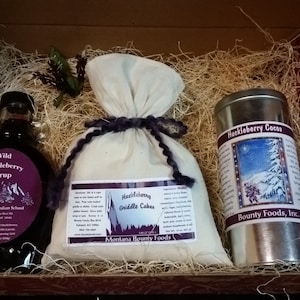 May include: A gift box filled with brown shredded paper contains a glass bottle of wild huckleberry syrup, a white bag of huckleberry griddle cakes mix, and a silver tin of huckleberry cocoa. The labels on the products feature a winter scene and the text "Montana Bounty Foods" and "Bounty Foods, Inc."