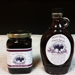 May include: Two glass jars with labels, one containing jam and the other syrup, both featuring a bison silhouette and the text "Montana Bounty Foods". The jam jar is labeled "Bison Berry Patch Jam" and the syrup jar is labeled "Bison Berry Patch Syrup".