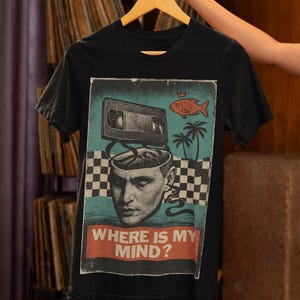 May include: Black t-shirt with a graphic design featuring a retro VHS tape, a fish, palm trees, and a man's head with the text "WHERE IS MY MIND?". The shirt is hanging on a wooden hanger.