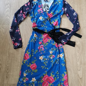 Puede incluir: Vestido cruzado azul con estampado floral, con rosas rosas y otras flores. Las mangas son de un azul más oscuro con diseño floral. Un cinturón negro ciñe la cintura. El vestido tiene un escote en V.