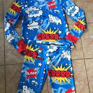 May include: A blue comic book-themed pajama set. The top and trousers are covered in action words such as "POW!" and "GRRRR!" in white speech bubbles and yellow starbursts. The long-sleeved top has red cuffs, and the trousers have a matching design.