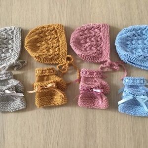 May include: Four sets of hand-knitted bonnets and matching booties in grey, mustard yellow, dusty pink, and light blue. Each bonnet features a textured cable knit pattern and side ties, while the booties include a ribbon bow detail.